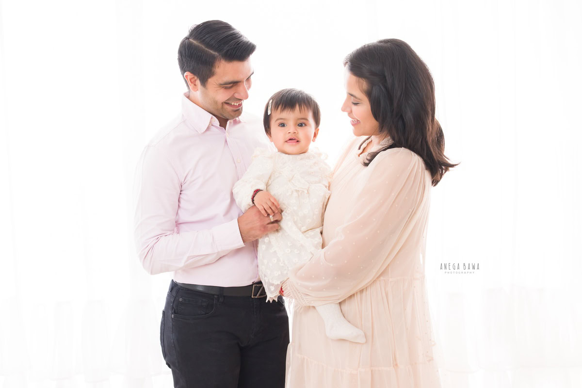 1-year-old girl posing cutely with mom and dad against a white backdrop, captured by Anega Bawa Photography for a family photoshoot in Gurgaon (Delhi NCR)