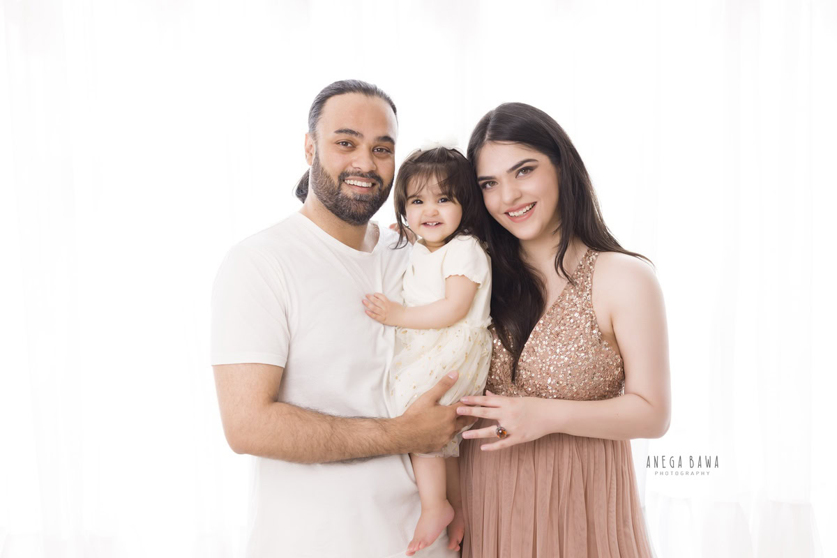 1-year-old girl posing cutely with mom and dad against a white backdrop, captured by Anega Bawa Photography for a family photoshoot in Gurgaon, Delhi NCR.