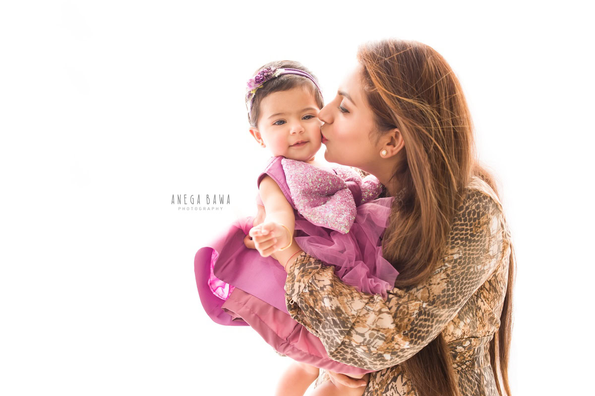 1-year-old girl posing cutely with mom in a kissing pose against a serene white backdrop, captured by Anega Bawa, family photographer in Gurgaon (Delhi NCR).