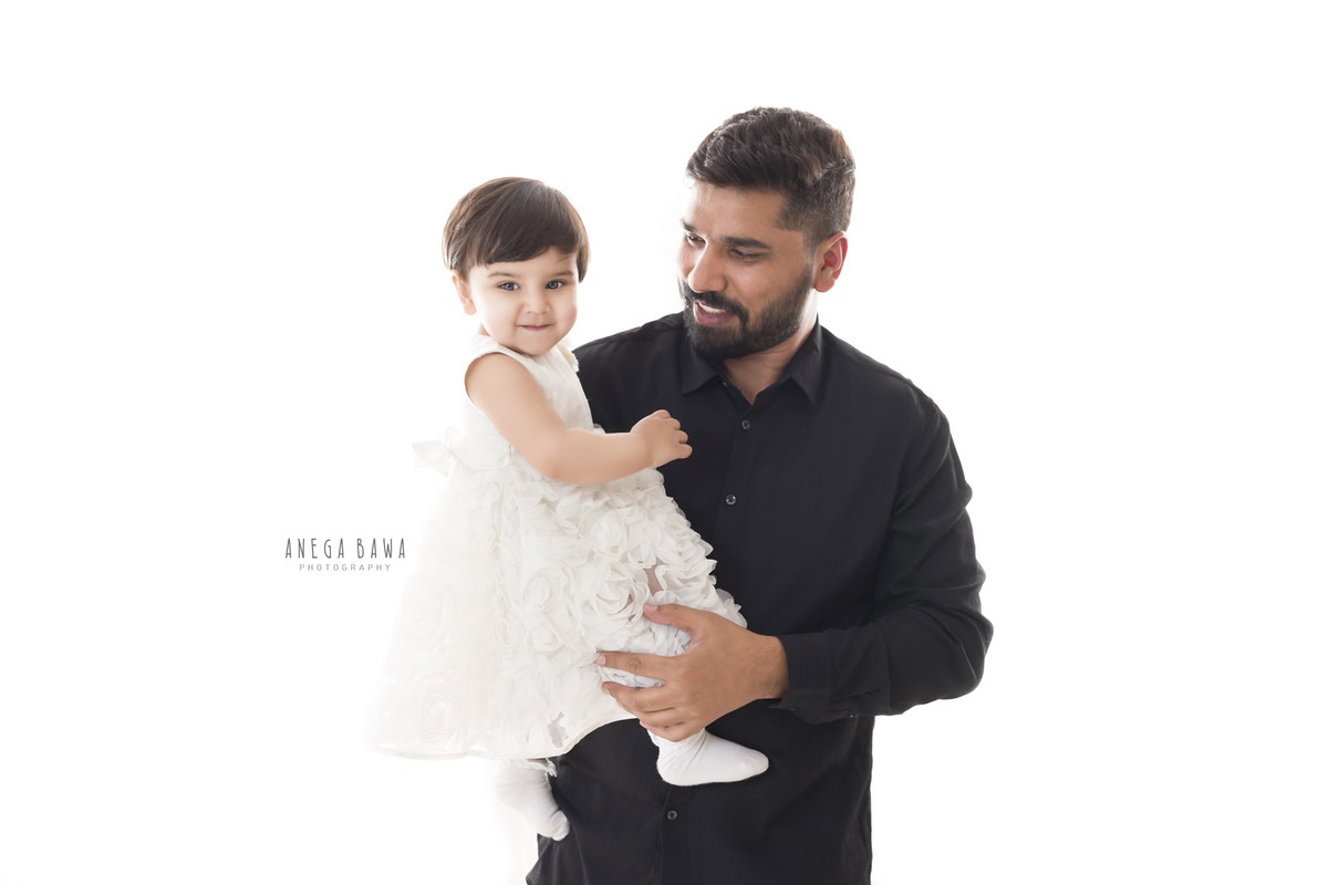 1-year-old girl posing in dad's arms against a white backdrop, captured by Anega Bawa, family photographer in Gurgaon (Delhi NCR).