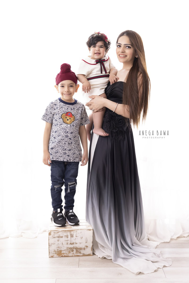 1-year girl posing in mom's arms with her elder brother against a white backdrop, captured by Anega Bawa, family photographer in Gurgaon (Delhi NCR).