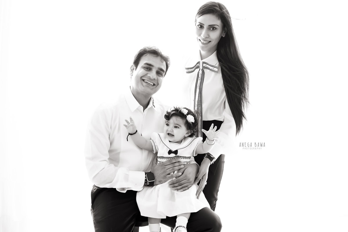 1-year-old girl posing on dad's lap with mom standing beside against a white backdrop, captured by Anega Bawa Photography for a family photoshoot in Gurgaon, Delhi NCR.