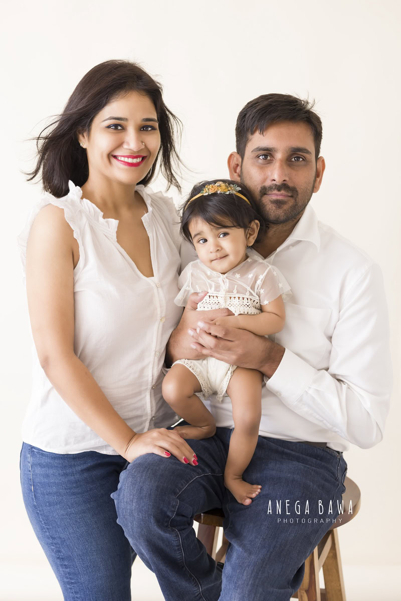 1-year-old girl posing on dad's lap with mom against a white backdrop, captured by Anega Bawa Family Photographer Gurgaon (Delhi NCR).