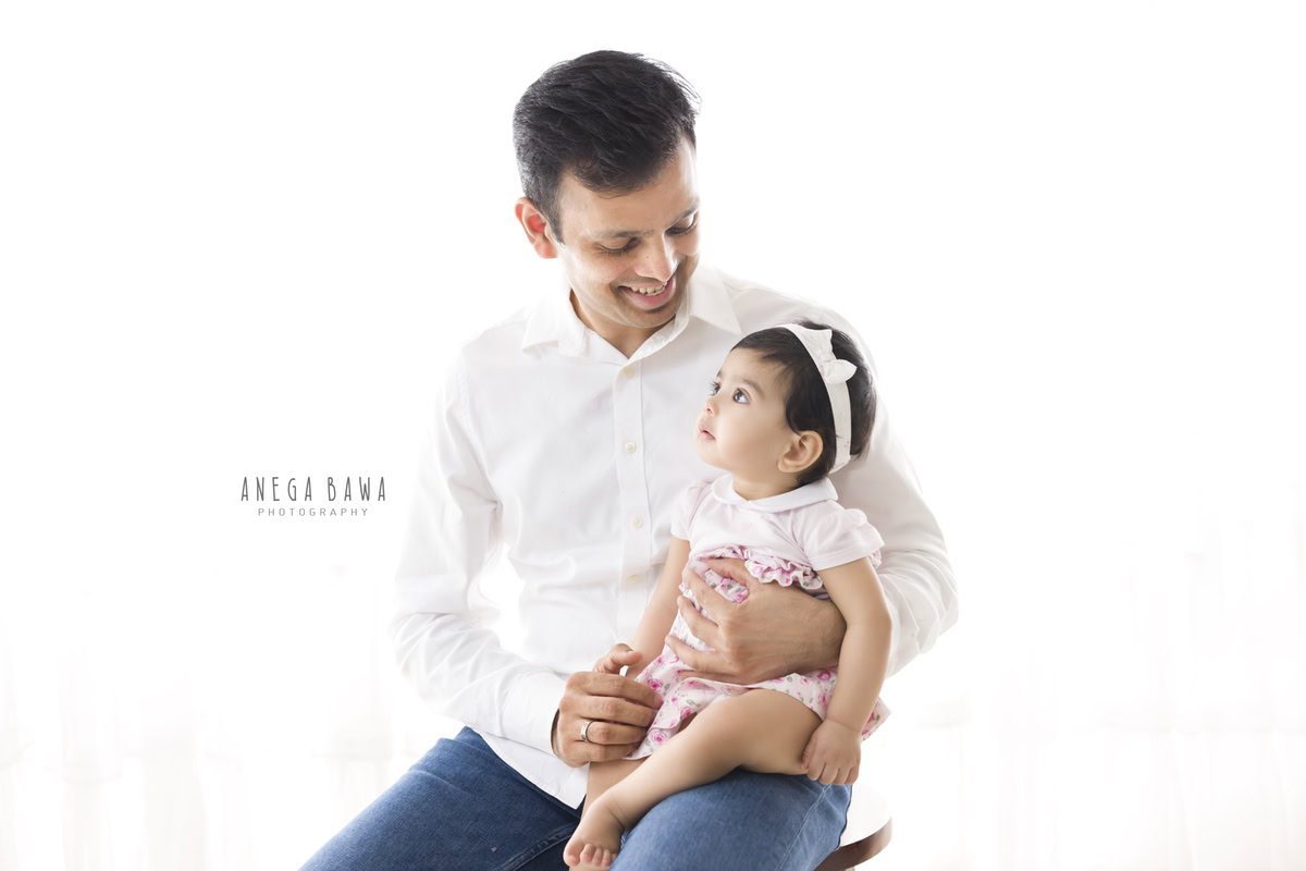 1-year-old girl posing on dad's lap against a white backdrop, captured by Anega Bawa Family Photographer Gurgaon (Delhi NCR).