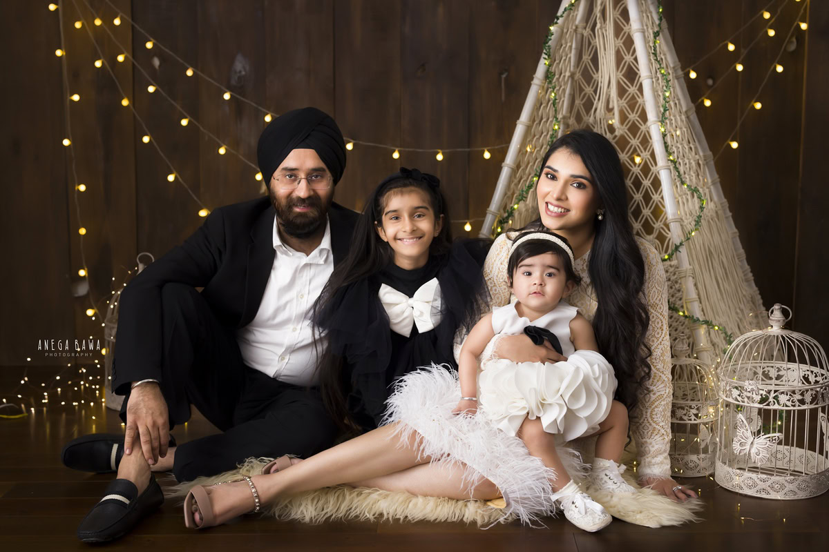 1-year-old girl posing on mom's lap with her elder sister and dad, surrounded by fairy lights and castles on a brown backdrop.