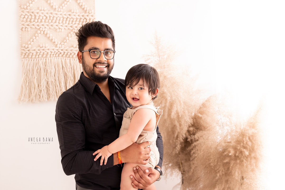 Here's the alt-text 1-year-old girl posing with dad against a backdrop of bushes and white backdrop, captured by Anega Bawa, family photographer in Gurgaon (Delhi NCR).