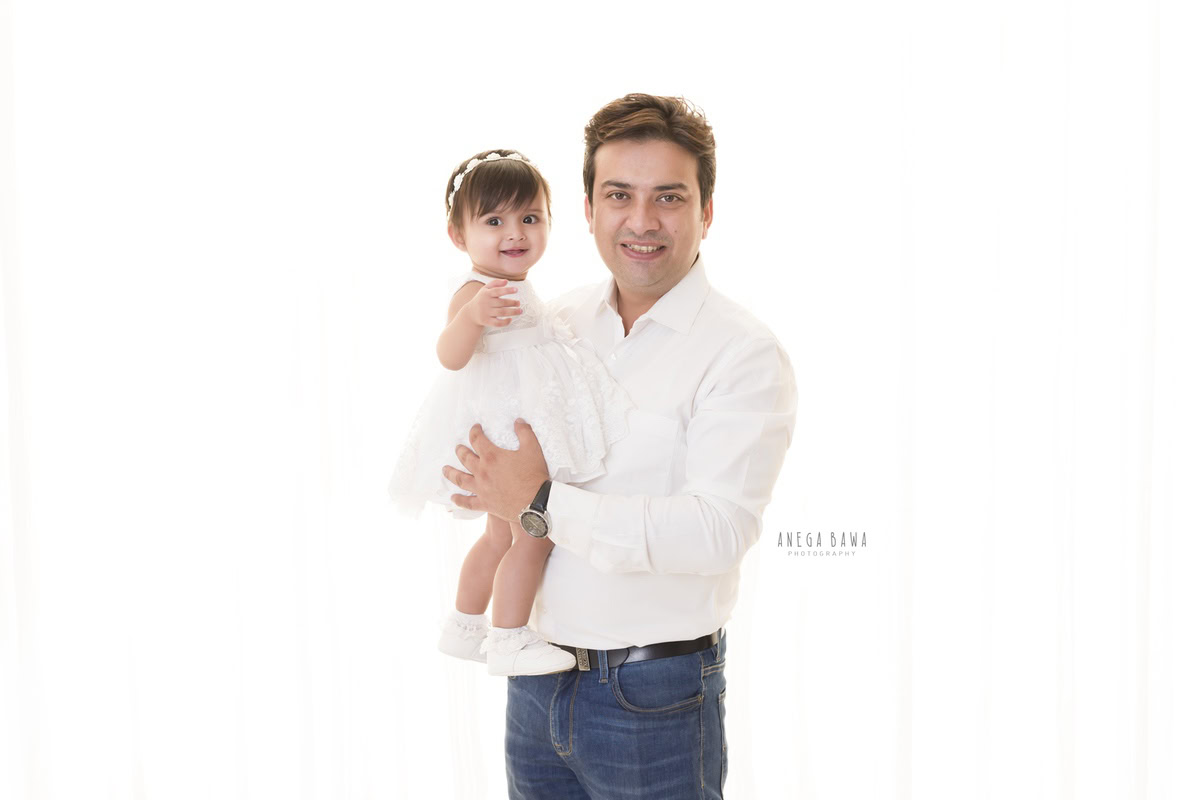 1-year-old girl posing with dad in a cute headband and white dress against a white backdrop, captured beautifully by Anega Bawa Photography for a family photoshoot in Gurgaon, Delhi NCR.