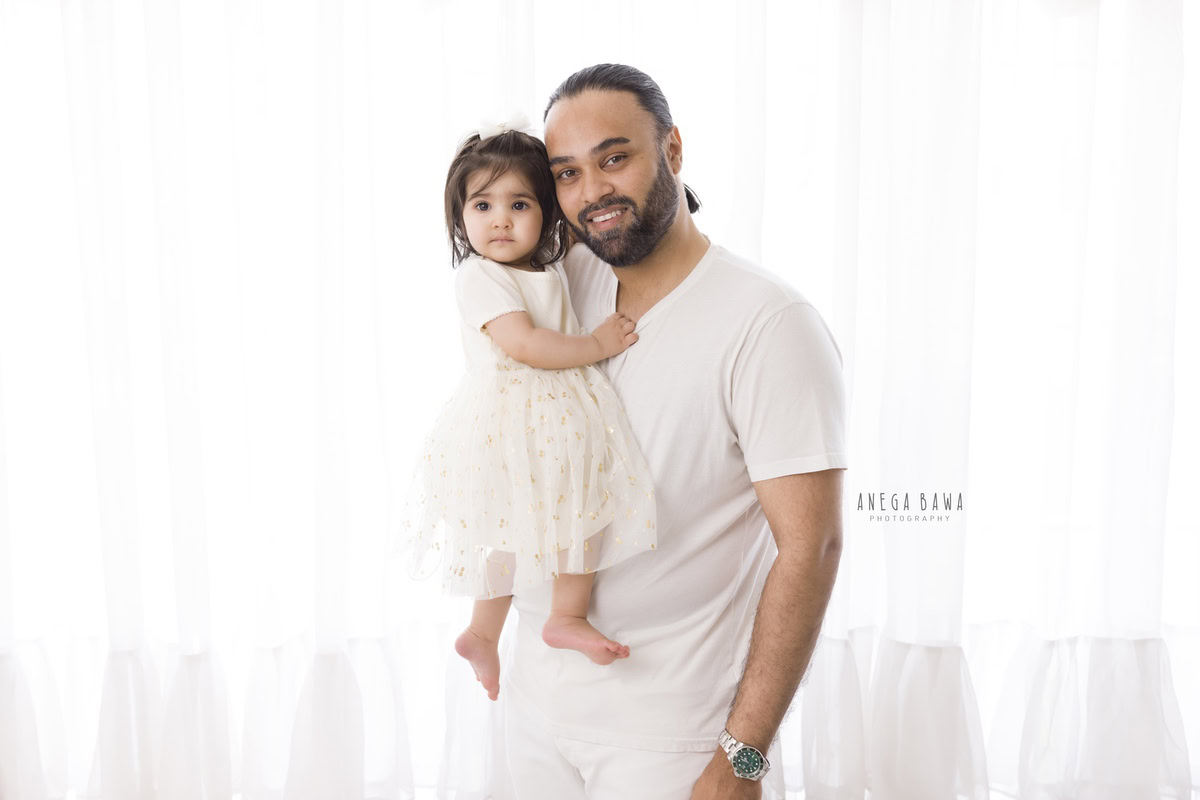 1-year-old girl posing with dad against a white backdrop, captured by Anega Bawa Family Photographer Gurgaon (Delhi NCR).