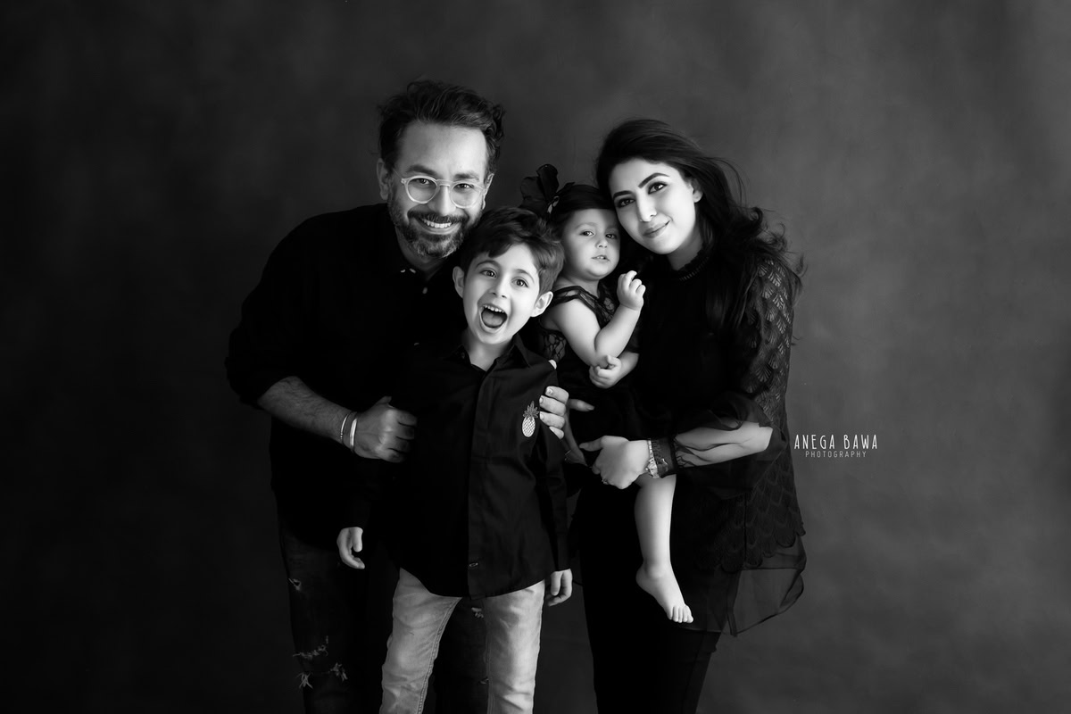 1-year-old girl posing with her elder brother, mom, and dad in a family pose against a black and white backdrop, all wearing black, captured by Anega Bawa Family Photographer Gurgaon (Delhi NCR).