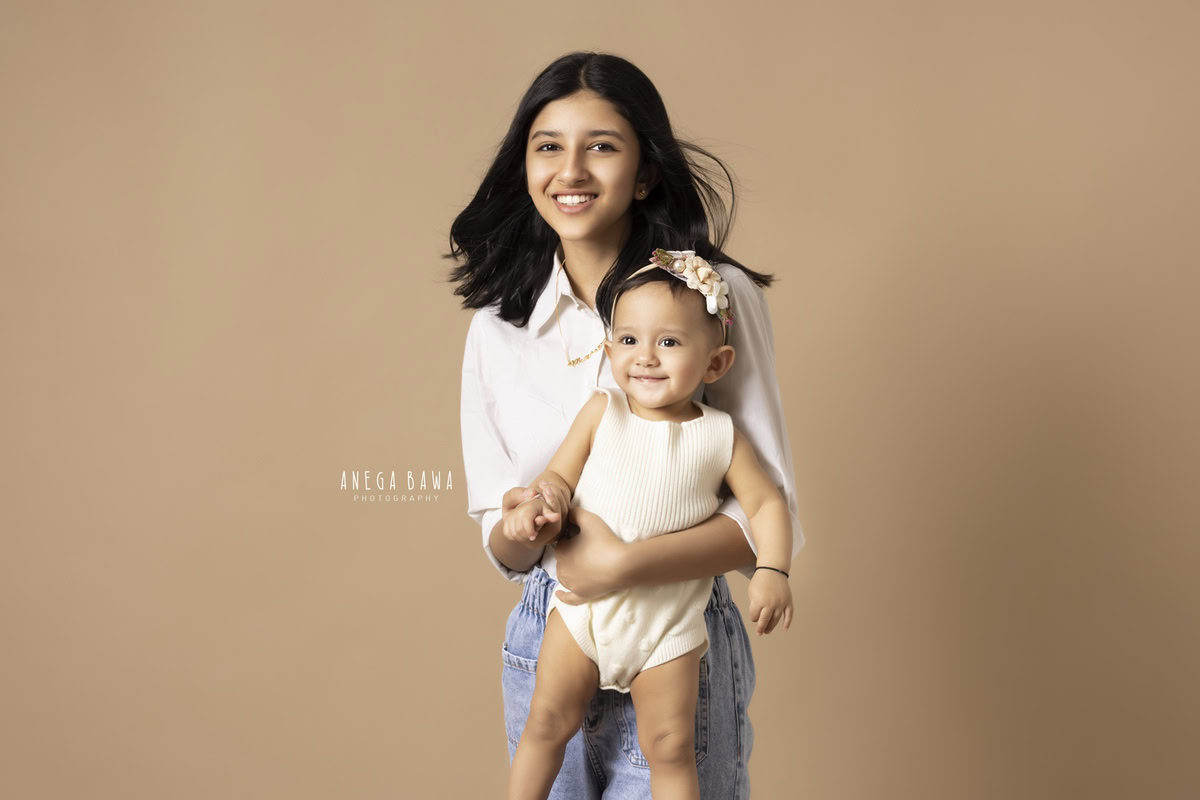 1-year-old girl posing with her elder sister against a light brown backdrop, captured by Anega Bawa Photography for a family photoshoot in Gurgaon (Delhi NCR).