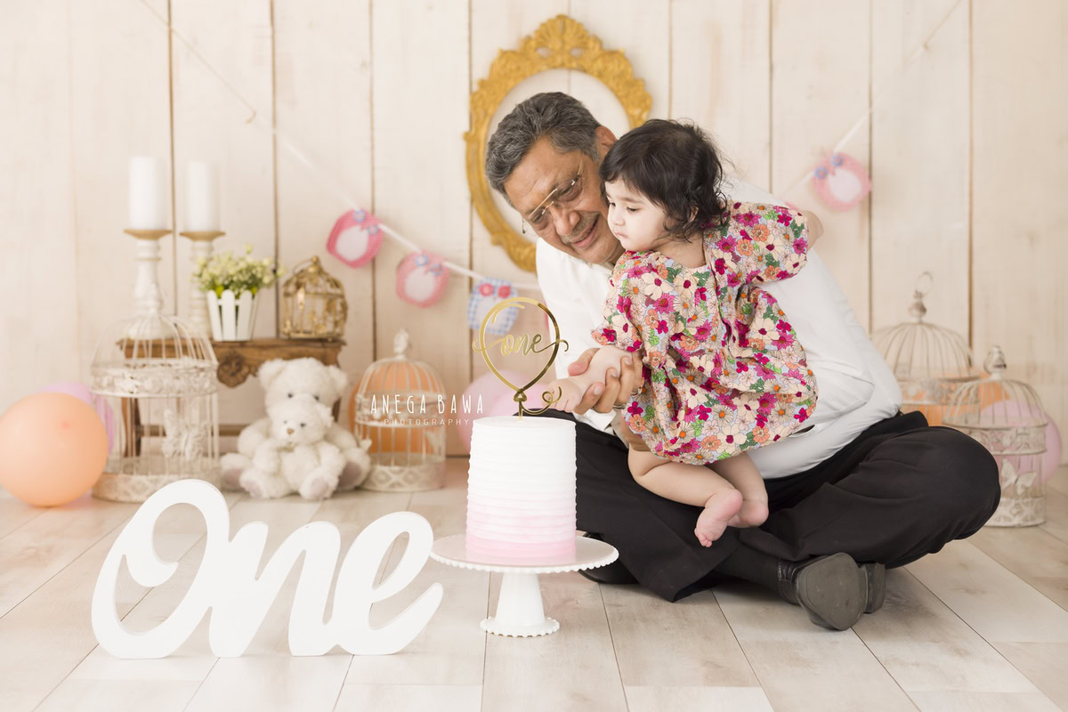 1-year-old girl posing with grandpa, cake, candlestand, and castles against a beige backdrop, captured by Anega Bawa Family Photographer Gurgaon (Delhi NCR).