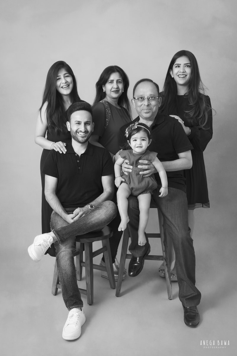1-year-old girl posing with grandparents, all wearing black, in a family pose against a grey backdrop. Captured by Anega Bawa Photography for a family photoshoot in Gurgaon (Delhi NCR).