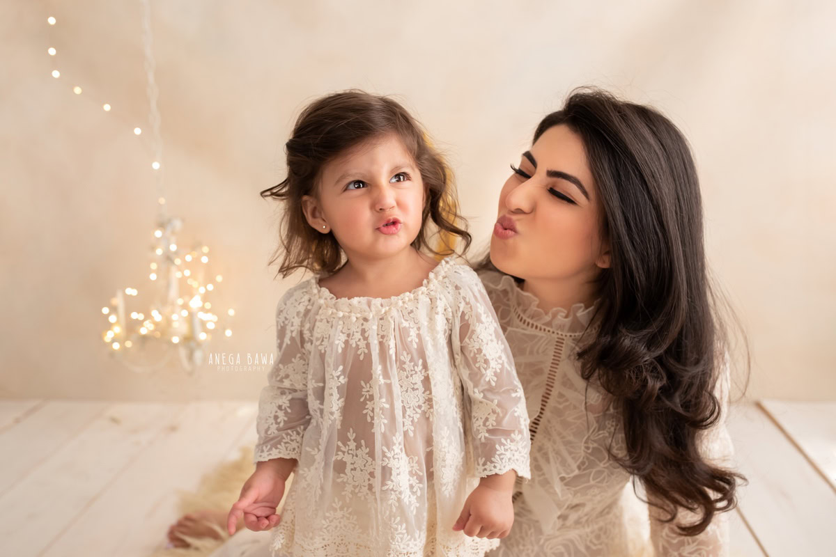 1-year-old girl posing with mom in a cute pout pose near a candle stand and fairy lights against a beige backdrop, captured by Anega Bawa, family photographer in Gurgaon (Delhi NCR).