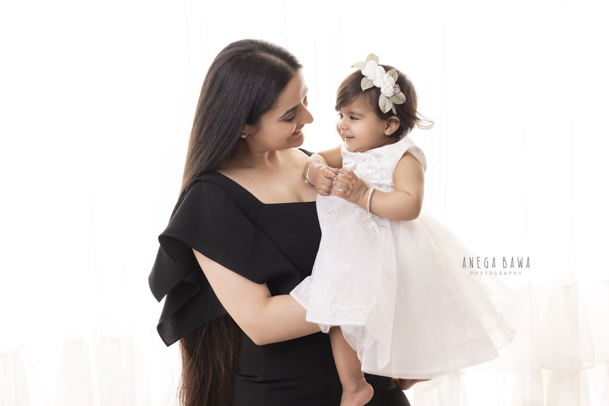 1-year-old girl posing with mom in a cute smiling pose against a white backdrop. Captured by Anega Bawa Photography for a family photoshoot in Gurgaon (Delhi NCR).