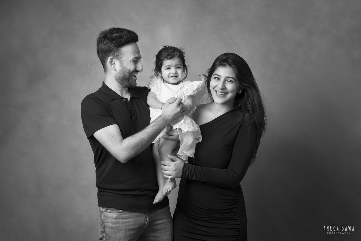 1-year girl posing with mom and dad in a black and white, smiling pose against a grey backdrop, captured by Anega Bawa Photography in Gurgaon (Delhi NCR).