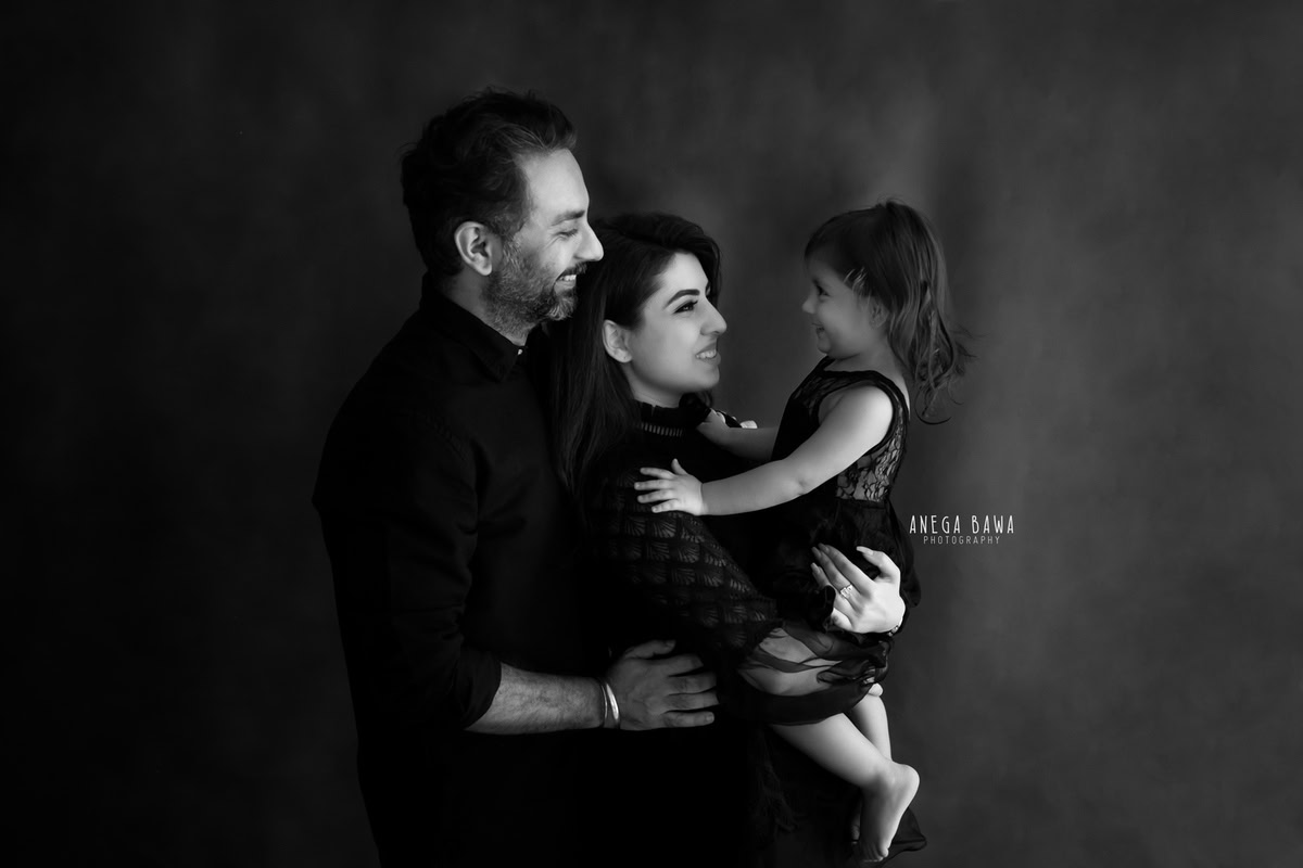 1-year-old girl posing with mom and dad against a black backdrop, all wearing black attire, captured by Anega Bawa, family photographer in Gurgaon (Delhi NCR).