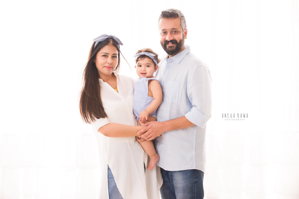 1-year girl posing with mom and dad, adorned with a cute headband, against a serene white backdrop, captured by Anega Bawa Photography in Gurgaon (Delhi NCR).