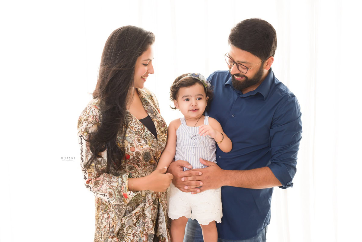 1-year girl posing with mom and dad, showcasing a cute smile against a white backdrop, captured by Anega Bawa Photography in Gurgaon (Delhi NCR).