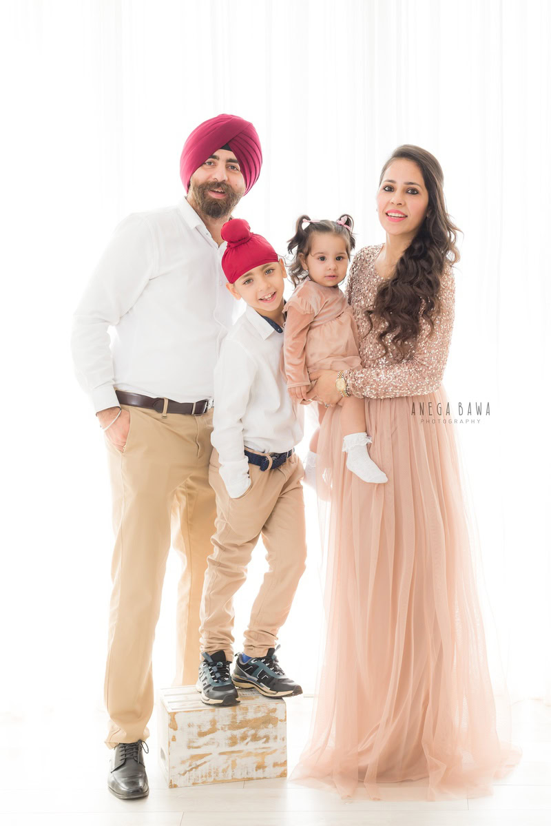 1-year-old girl posing with mom, dad, and elder brother, all in white outfits, against a beige and white backdrop, captured by Anega Bawa Family Photographer Gurgaon (Delhi NCR).