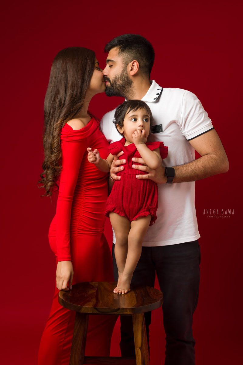 1-year-old girl posing with mom and dad in a kissing pose against a red backdrop, family photoshoot by Anega Bawa, Gurgaon (Delhi, NCR)