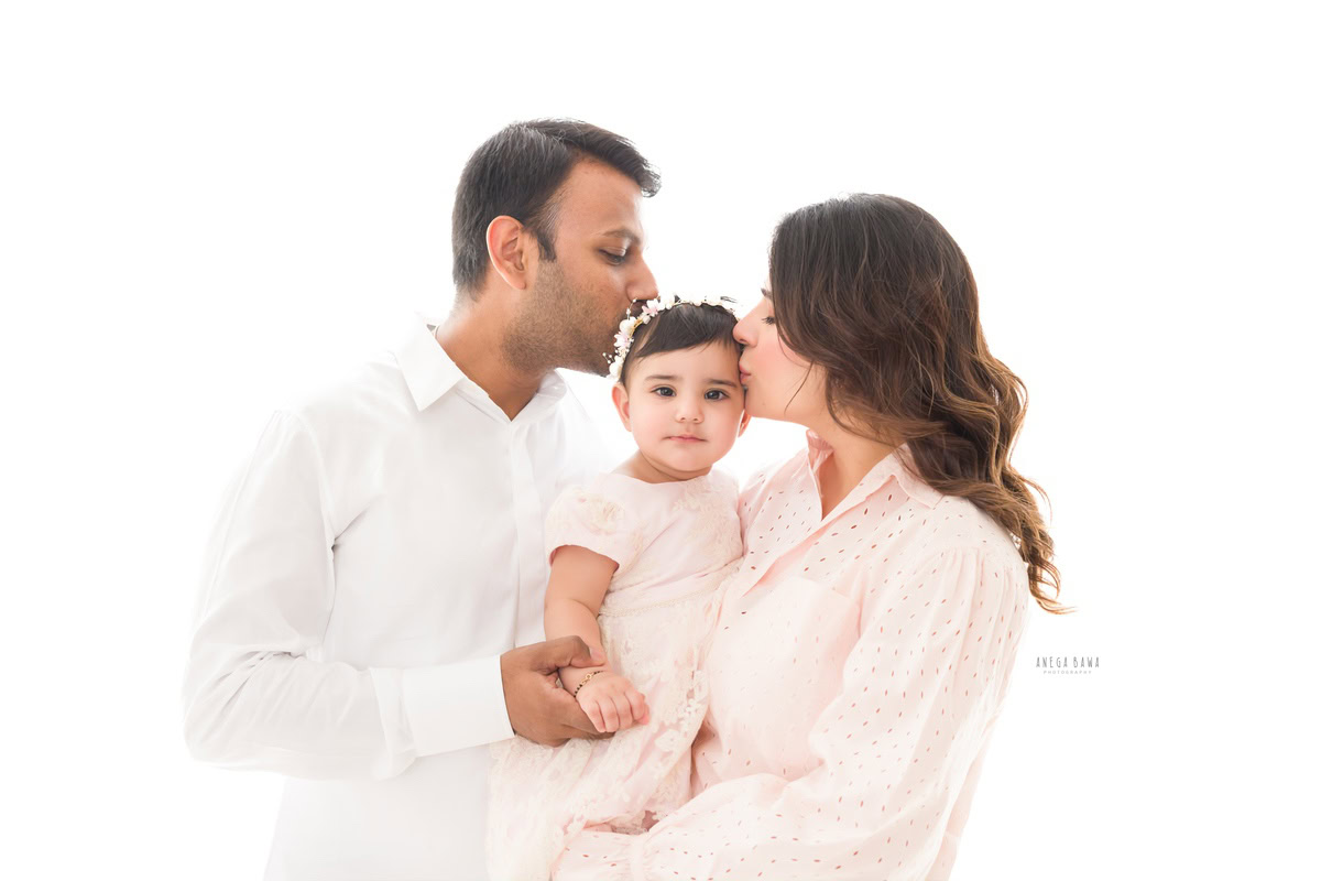 1-year girl posing with mom and dad in a tender kissing pose against a serene white backdrop, captured beautifully by Anega Bawa Photography in Gurgaon (Delhi NCR).