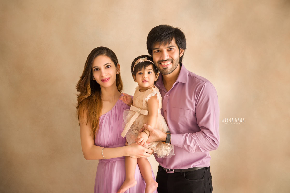 1-year-old girl posing with mom and dad against a light brown backdrop. Captured beautifully by Anega Bawa Photography for a family photoshoot in Gurgaon (Delhi NCR).