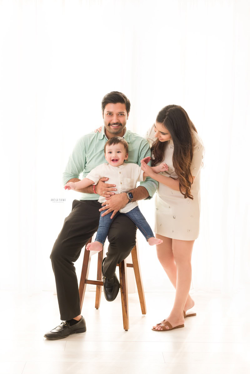 1-year girl posing with mom and dad seated on a wooden stool against a clean white backdrop, captured by Anega Bawa Photography in Gurgaon (Delhi NCR).