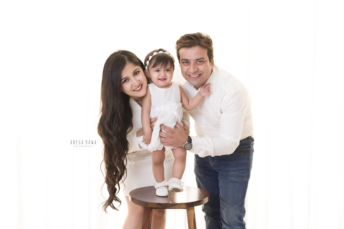 1-year-old girl posing with mom and dad, standing on a stool, dressed in a cute white dress and headband, against a white backdrop, captured by Anega Bawa Family Photographer Gurgaon (Delhi NCR).