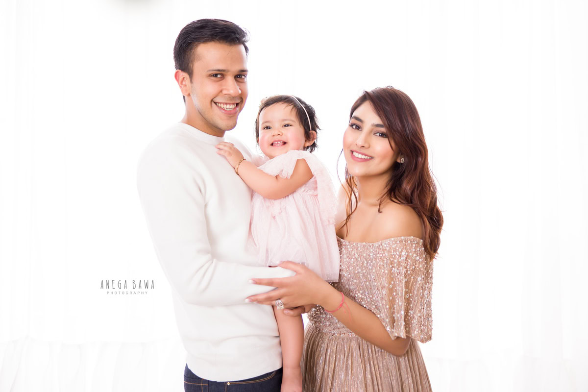 1-year-old girl posing with mom and dad in front of a white backdrop, photographed by Anega Bawa Photography in Gurgaon (Delhi NCR).