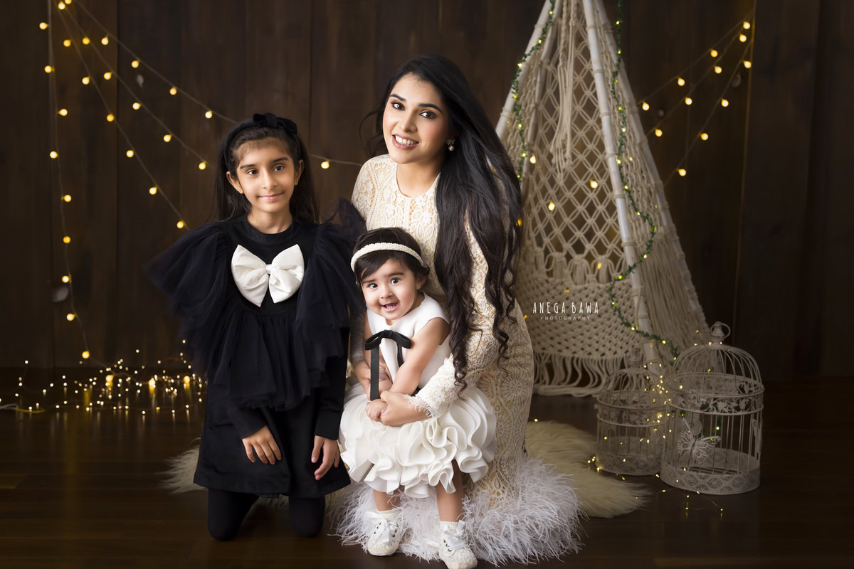1-year-old girl posing with her mom and elder sister, seated gracefully on a beige rug surrounded by fairy lights against a serene brown backdrop. Captured by Anega Bawa Photography, renowned for family photoshoots in Gurgaon (Delhi NCR), this moment captures the essence of familial joy and warmth.