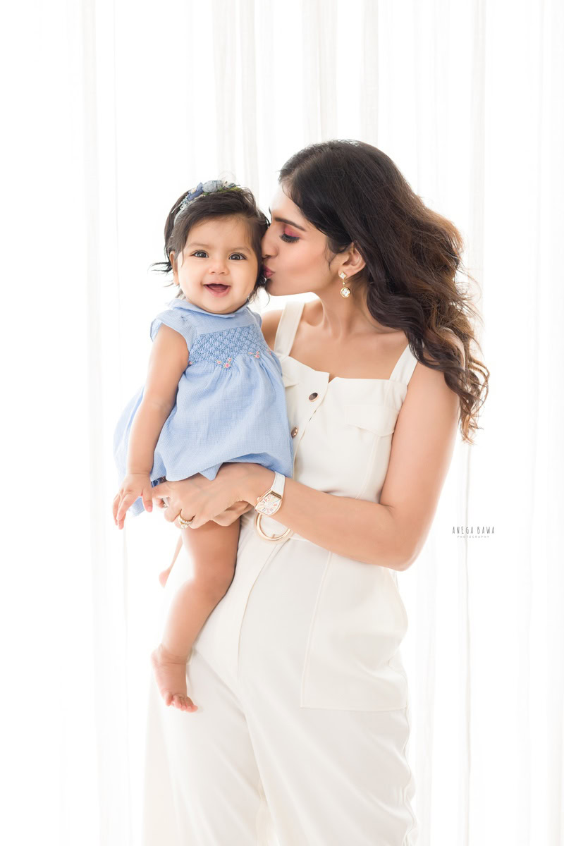 1-year-old girl posing with mom in a kissing pose against a white backdrop, captured by Anega Bawa, family photographer in Gurgaon (Delhi NCR).