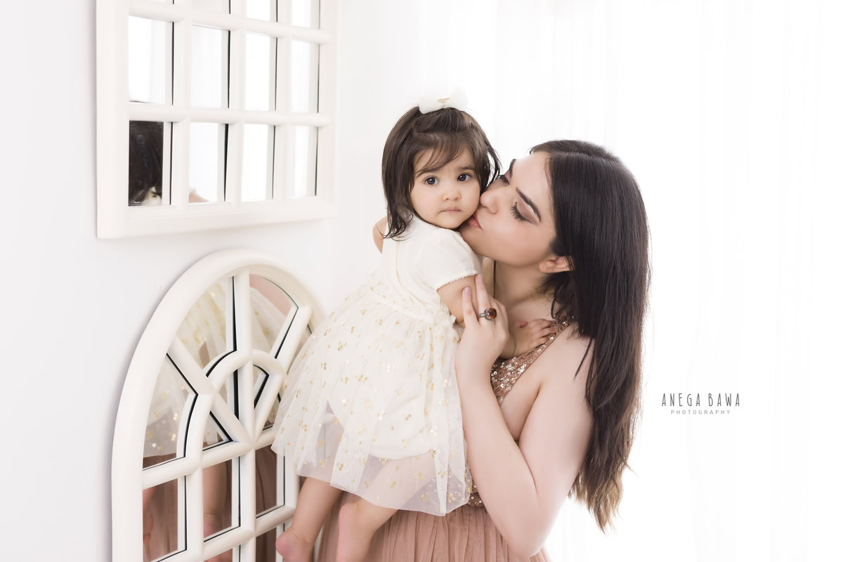 1-year-old girl posing with mom in a kissing pose against a backdrop with a white window, captured by Anega Bawa Family Photographer Gurgaon (Delhi NCR).