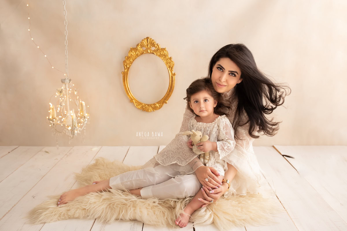 1-year-old girl posing with her mom, seated elegantly on a beige rug with a candle stand and golden frame on the wall, against a serene beige backdrop. Capture this beautiful moment with Anega Bawa Photography in Gurgaon (Delhi NCR), specializing in family photo shoots.