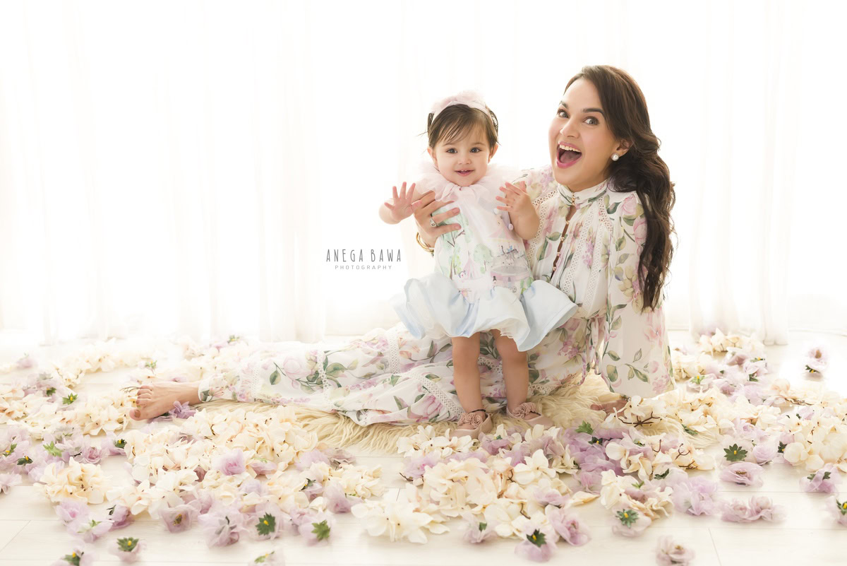 1-year-old girl posing with mom, seated on the floor with a beige rug and flowers, against a white backdrop, family photoshoot by Anega Bawa, Gurgaon (Delhi, NCR)