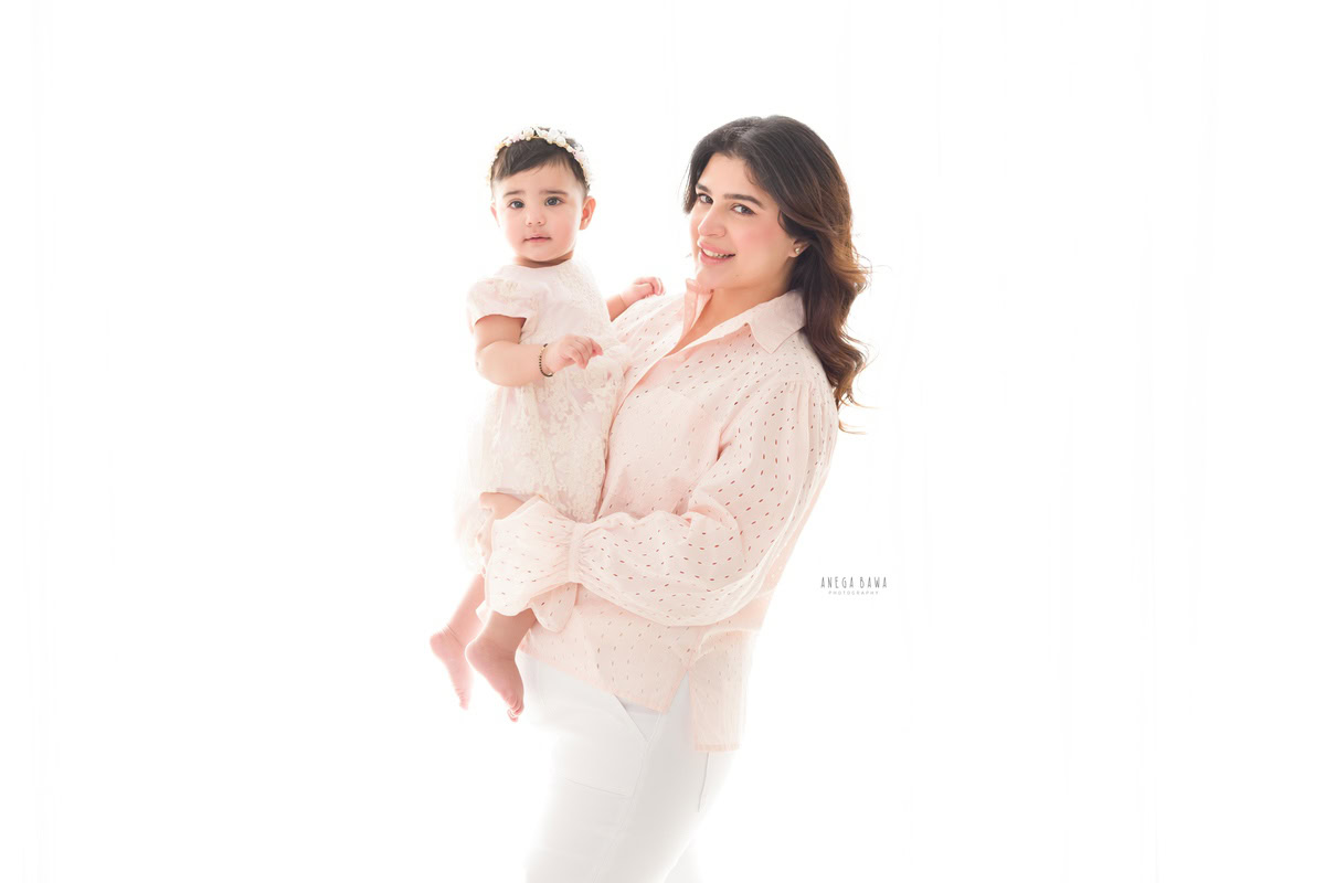 Adorable 1-year-old girl posing with her mom against a serene white backdrop. Captured by Anega Bawa Photography, this heartwarming moment is part of a family photo shoot in Gurgaon (Delhi NCR), showcasing the special bond between mother and daughter.