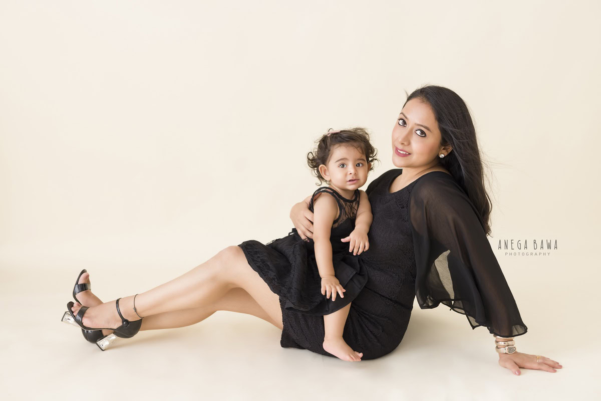 1-year-old girl sitting cutely on mom's lap against a beige backdrop, captured by Anega Bawa, family photographer in Gurgaon (Delhi NCR).