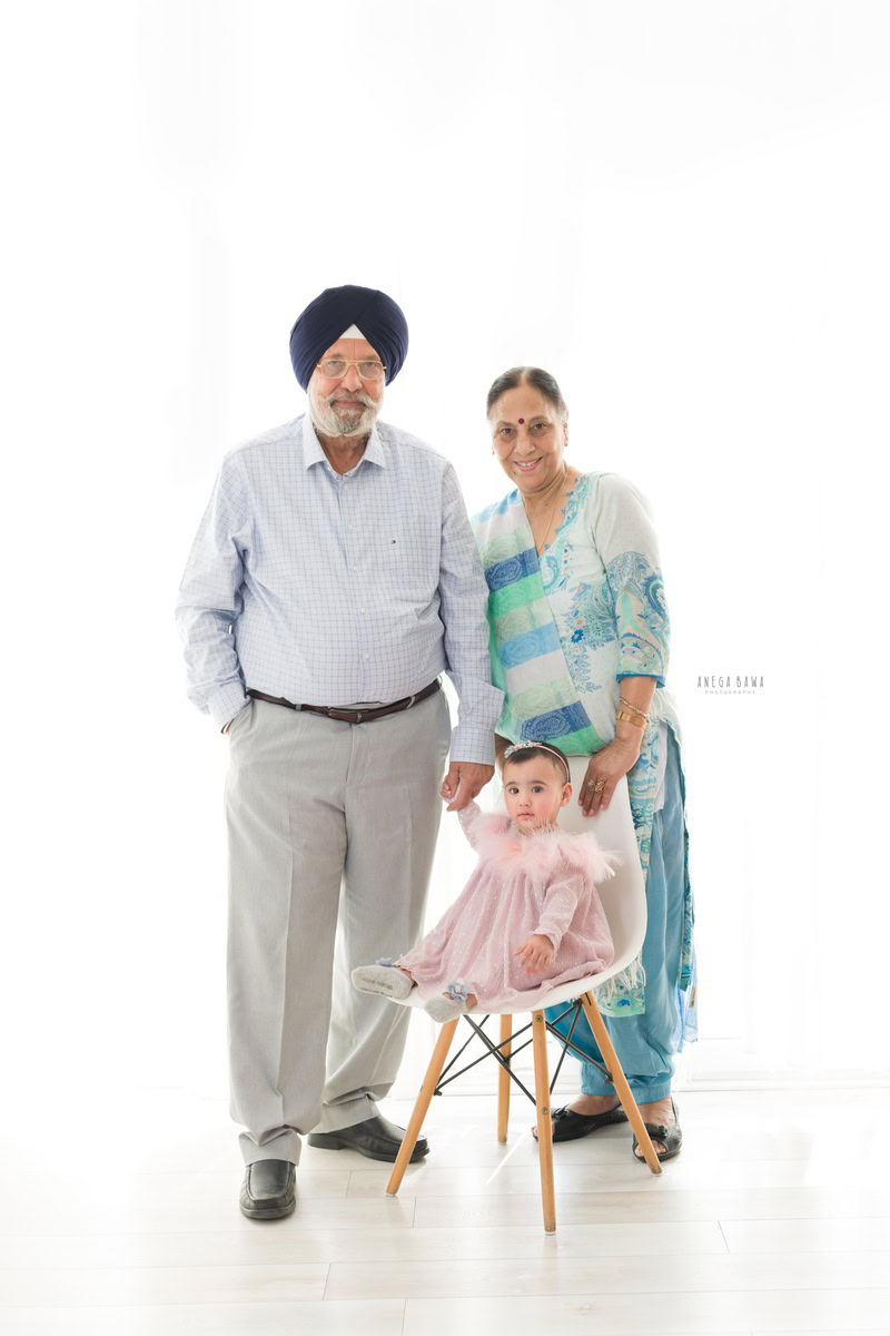 1-year-old girl seated on a chair, posing with her grandparents against a white backdrop, beautifully captured by Anega Bawa Photography in Gurgaon (Delhi NCR).