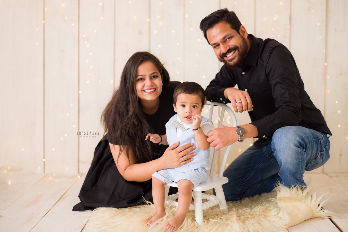 Sweet 1-year-old girl seated on a chair, posing with her mom and dad against a beige backdrop. Captured by Anega Bawa Photography, this charming family moment highlights the joy of togetherness in Gurgaon (Delhi NCR).