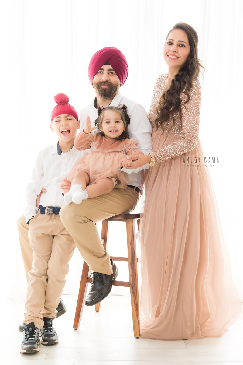 1-year-old girl seated on dad's lap in a cute smiling pose, with mom and elder brother, against a white backdrop, family photoshoot by Anega Bawa, Gurgaon (Delhi, NCR)