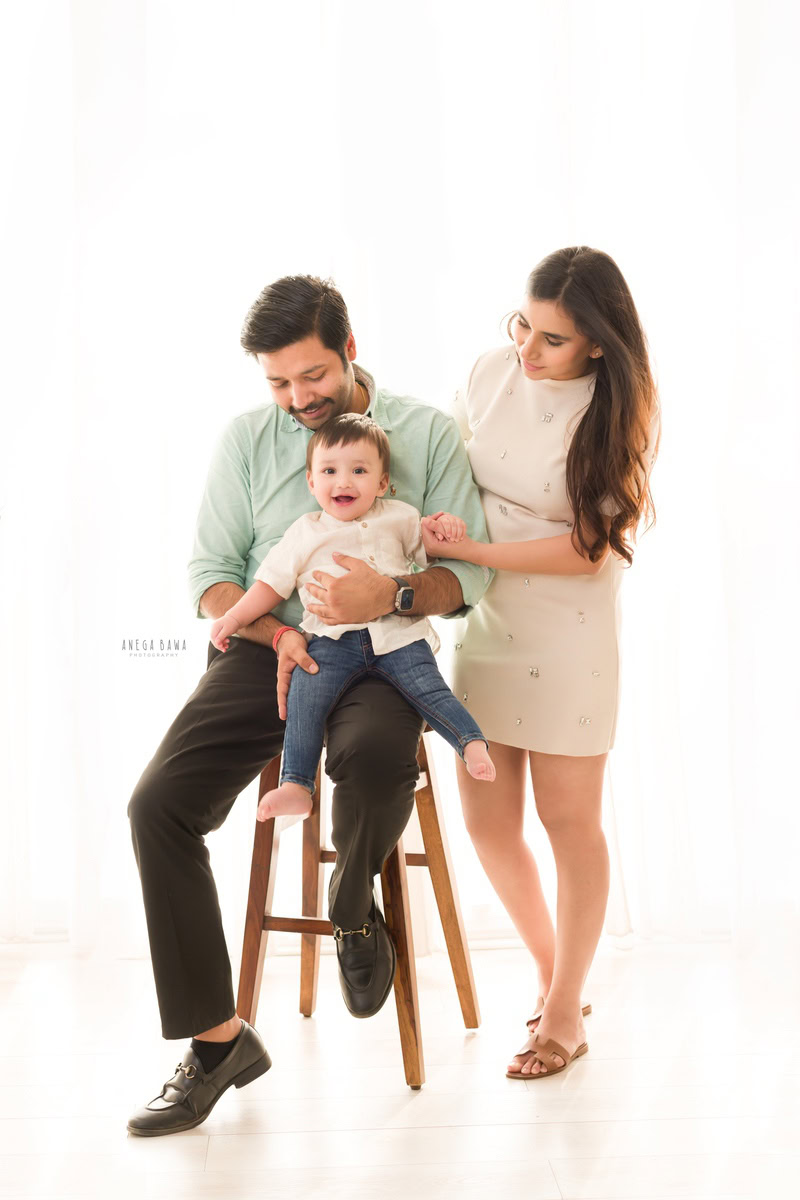 1-year-old girl sitting on dad's lap, cute and smiling with mom against a white backdrop, captured by Anega Bawa, family photographer in Gurgaon (Delhi NCR).