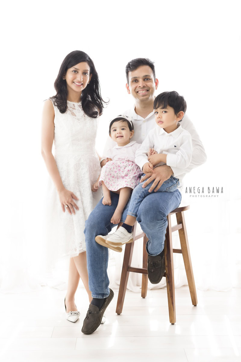 Adorable 1-year-old girl seated on dad's lap, with her elder brother and mom posing together in a heartwarming family pose against a serene white backdrop. Captured by Anega Bawa Photography, celebrating precious family moments in Gurgaon (Delhi NCR).