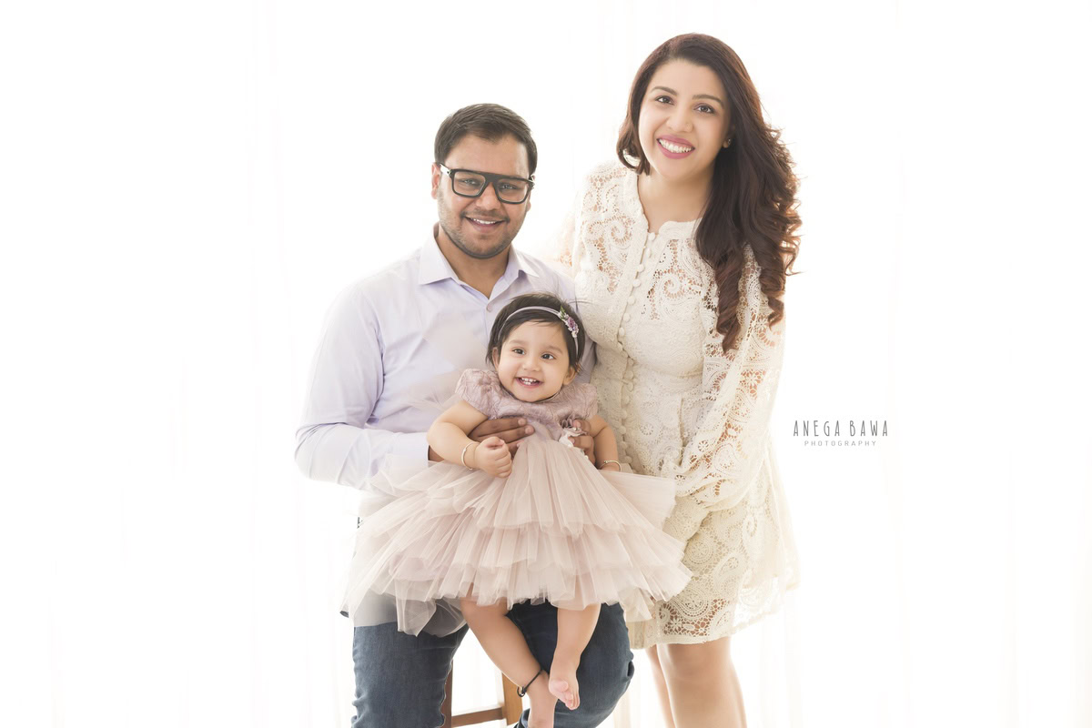 Heartwarming family photoshoot in Gurgaon: 1-year-old girl sitting on her dad's lap while mom poses against a white backdrop. Captured by Anega Bawa Photography, Gurgaon (Delhi, NCR).
