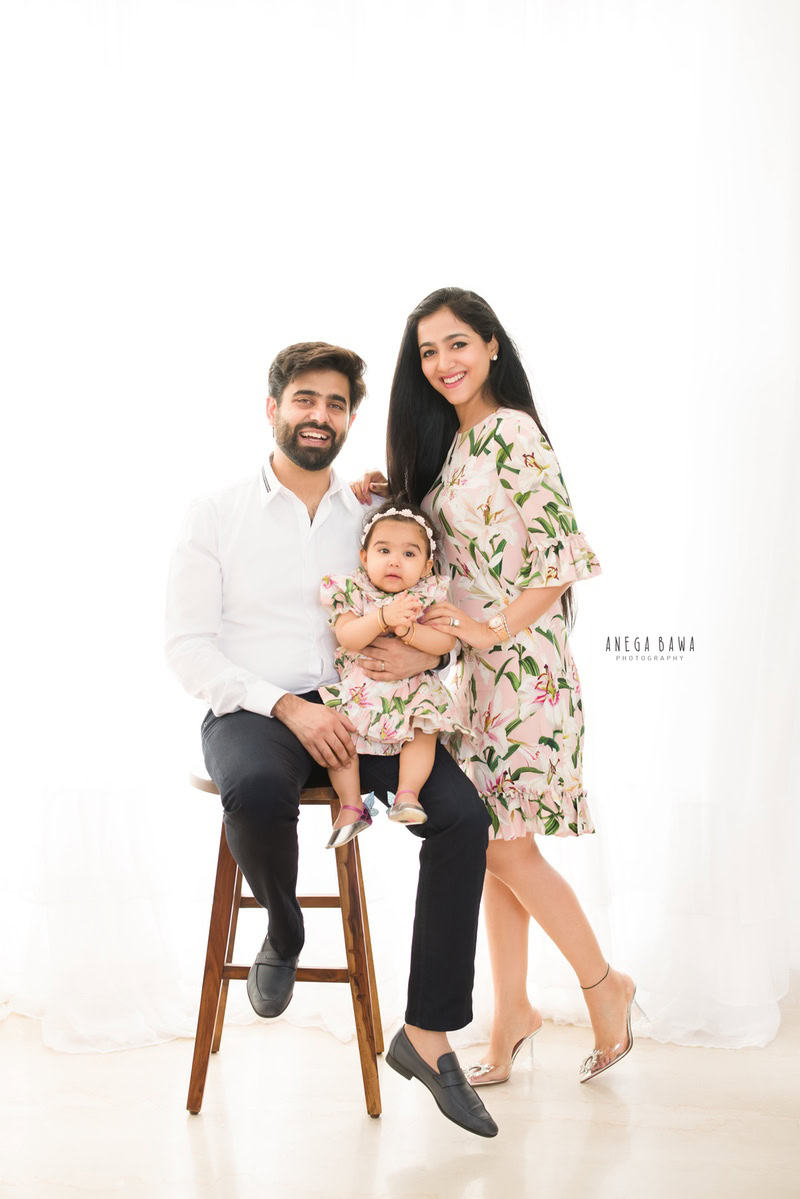 1-year-old girl seated on dad's lap, with mom posing, against a white backdrop, family photoshoot by Anega Bawa, Gurgaon (Delhi, NCR)