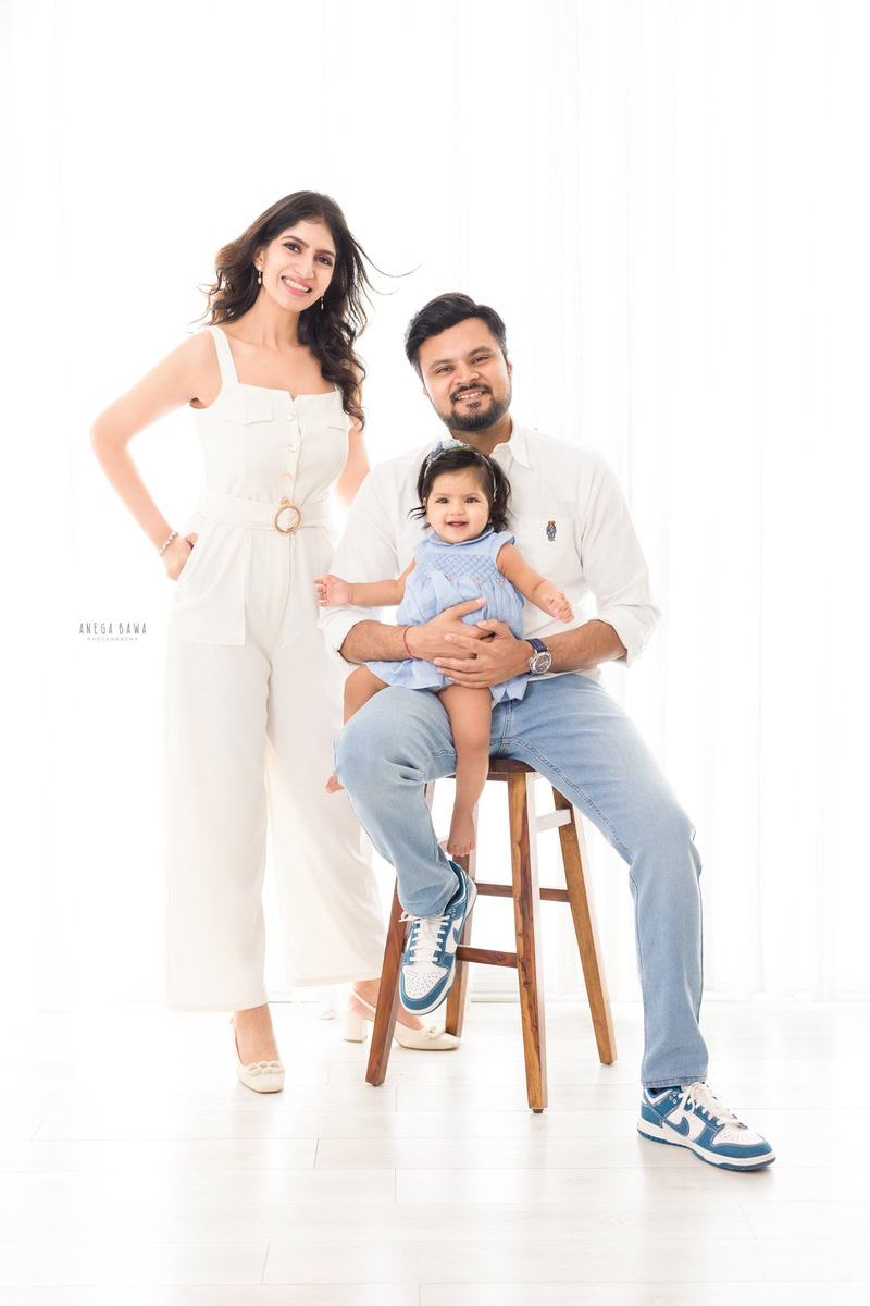 1-year-old girl seated on her dad's lap with mom standing beside them against a pristine white backdrop. Captured by Anega Bawa Photography, Gurgaon (Delhi, NCR).