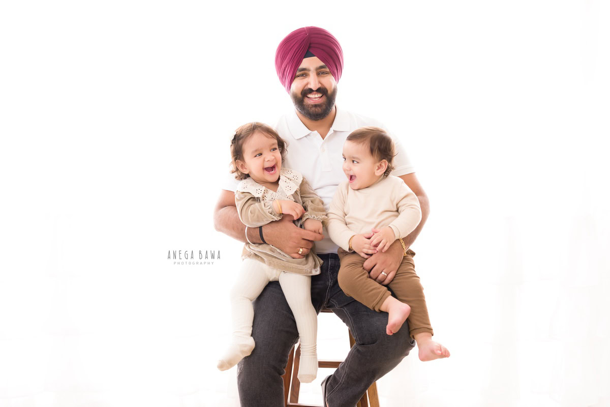 1-year-old girl seated on dad's lap, posing with elder brother against a white backdrop, captured by Anega Bawa, family photographer in Gurgaon (Delhi NCR).
