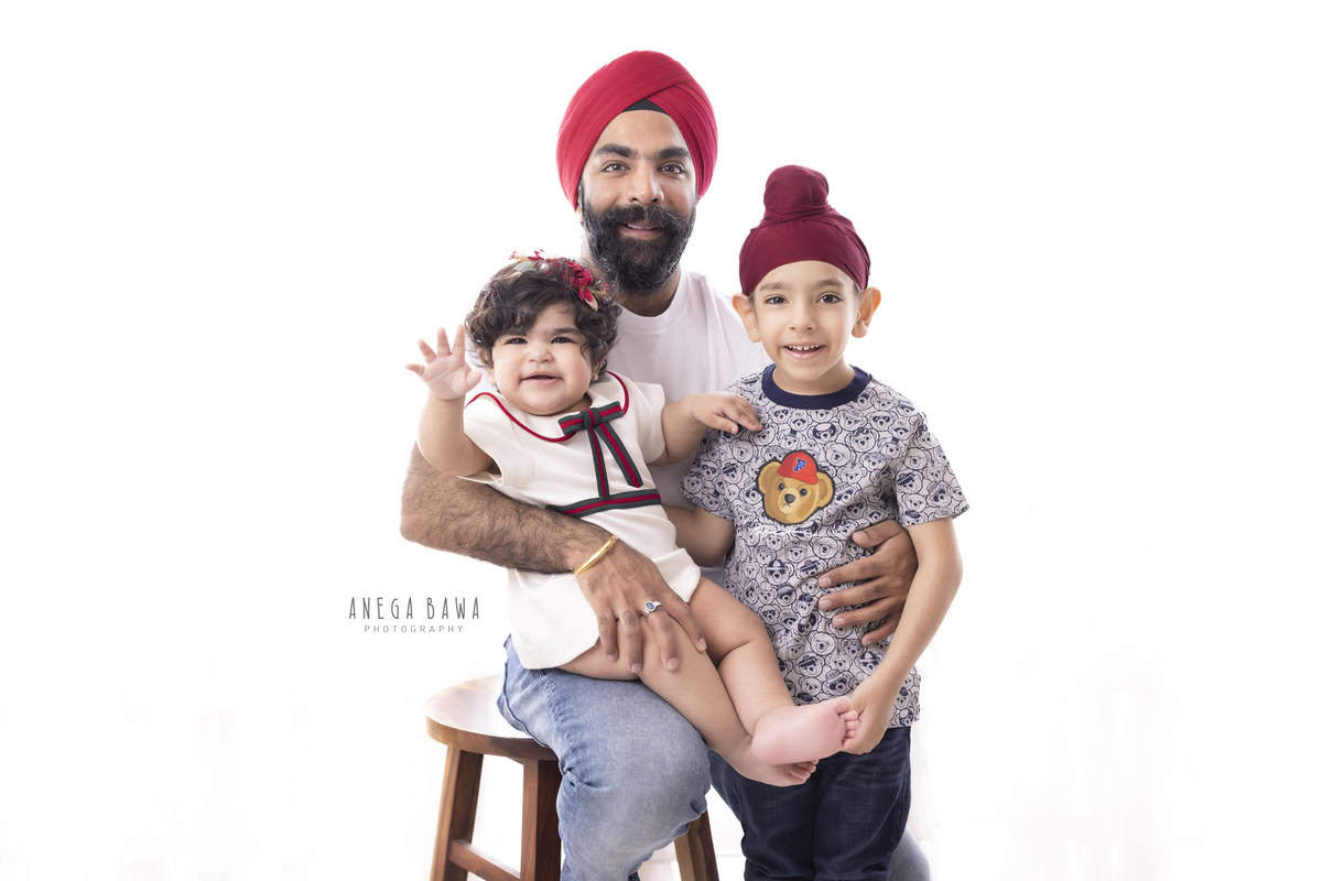 Sweet 1-year-old girl seated on dad's lap, posing with her elder brother against a clean white backdrop. This heartwarming family moment was captured by Anega Bawa Photography, specializing in capturing family memories in Gurgaon (Delhi NCR).