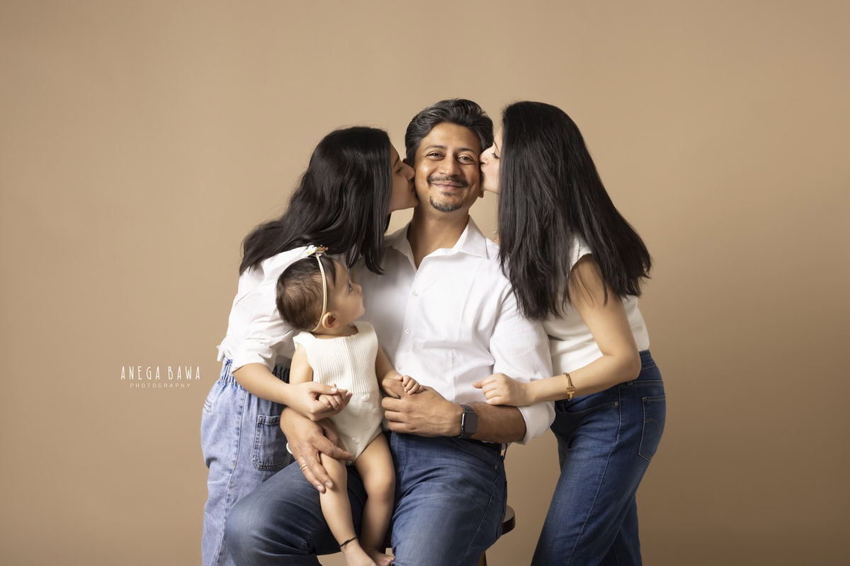 1-year-old girl seated on dad's lap, posing with her elder sister, while mom kisses dad, against a light brown backdrop. Captured by Anega Bawa Photography in Gurgaon (Delhi NCR).