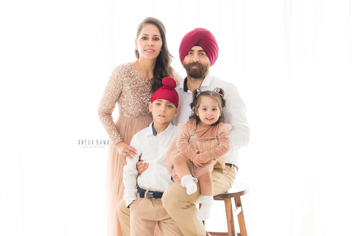 1-year-old girl seating on dad's lap, posing with mom and elder brother in a family pose against a white backdrop, captured by Anega Bawa, family photographer in Gurgaon (Delhi NCR).