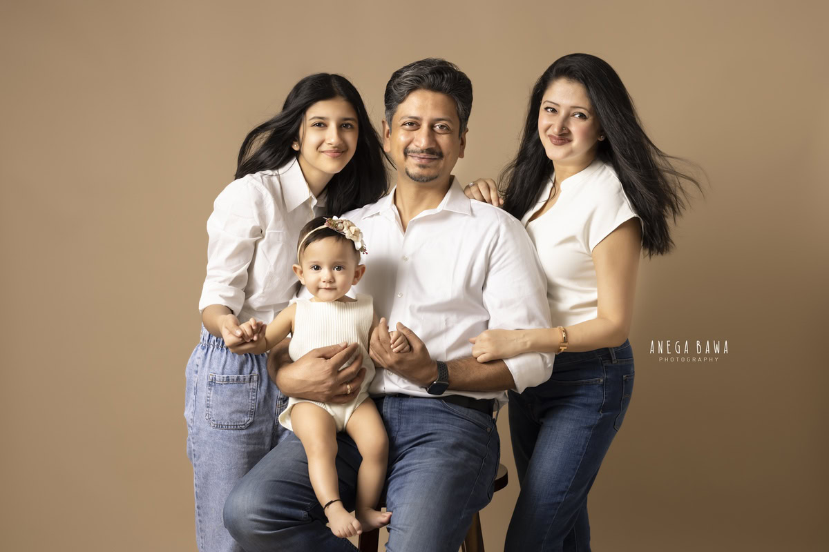1-year-old girl seated on dad's lap, posing with mom and elder sister in a family pose against a light brown backdrop, family photoshoot by Anega Bawa, Gurgaon (Delhi, NCR)