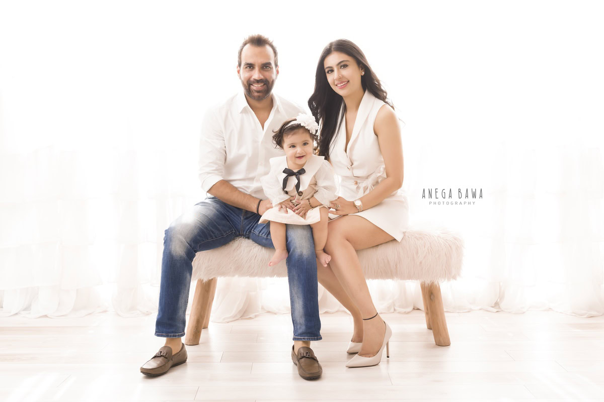 Adorable 1-year-old girl seated on dad's lap, posing with mom seated on a couch against a serene white backdrop. Experience the warmth of family moments captured by Anega Bawa Photography in Gurgaon (Delhi NCR).